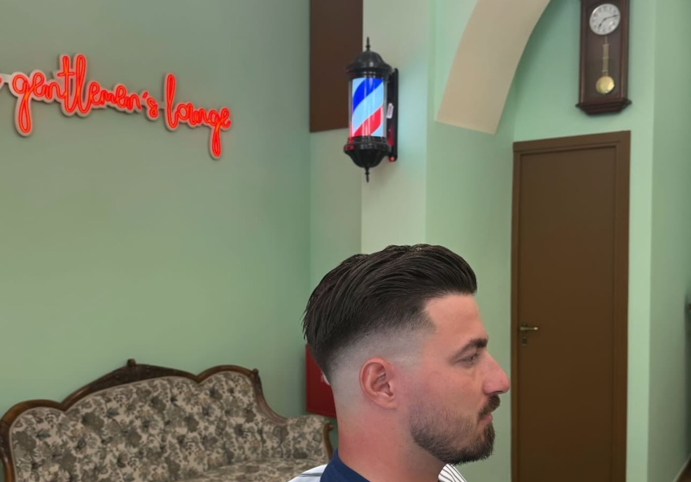Το εσωτερικό του The Soprano’s Barbershop στην Κάτω Τούμπα: η ρετρό πολυθρόνα, το εκκρεμές ρολόι και το κόκκινο νέον